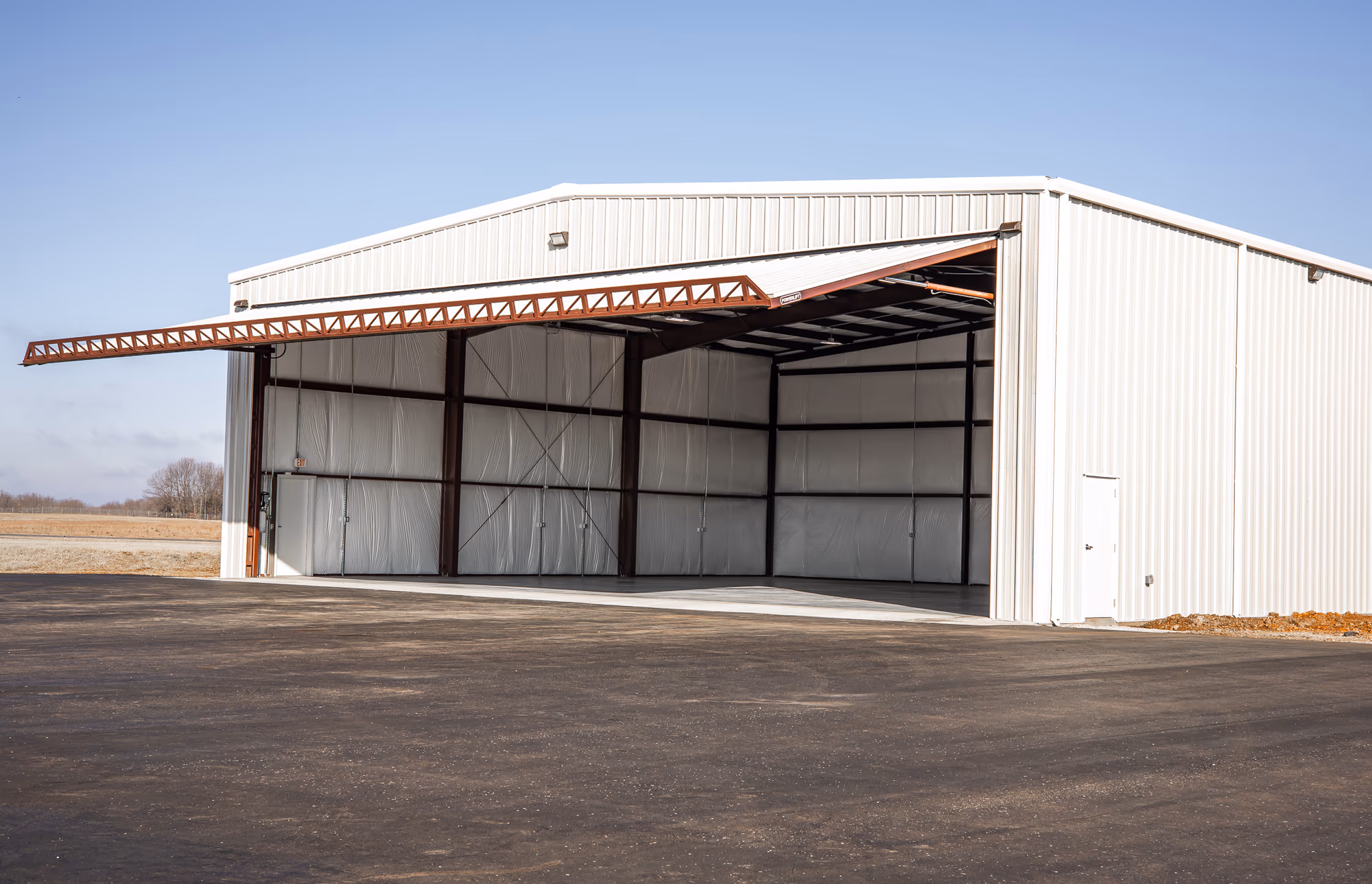 A white steel aircraft hangar with its full-width hydraulic bifold door raised on a red-iron frame, open to the apron