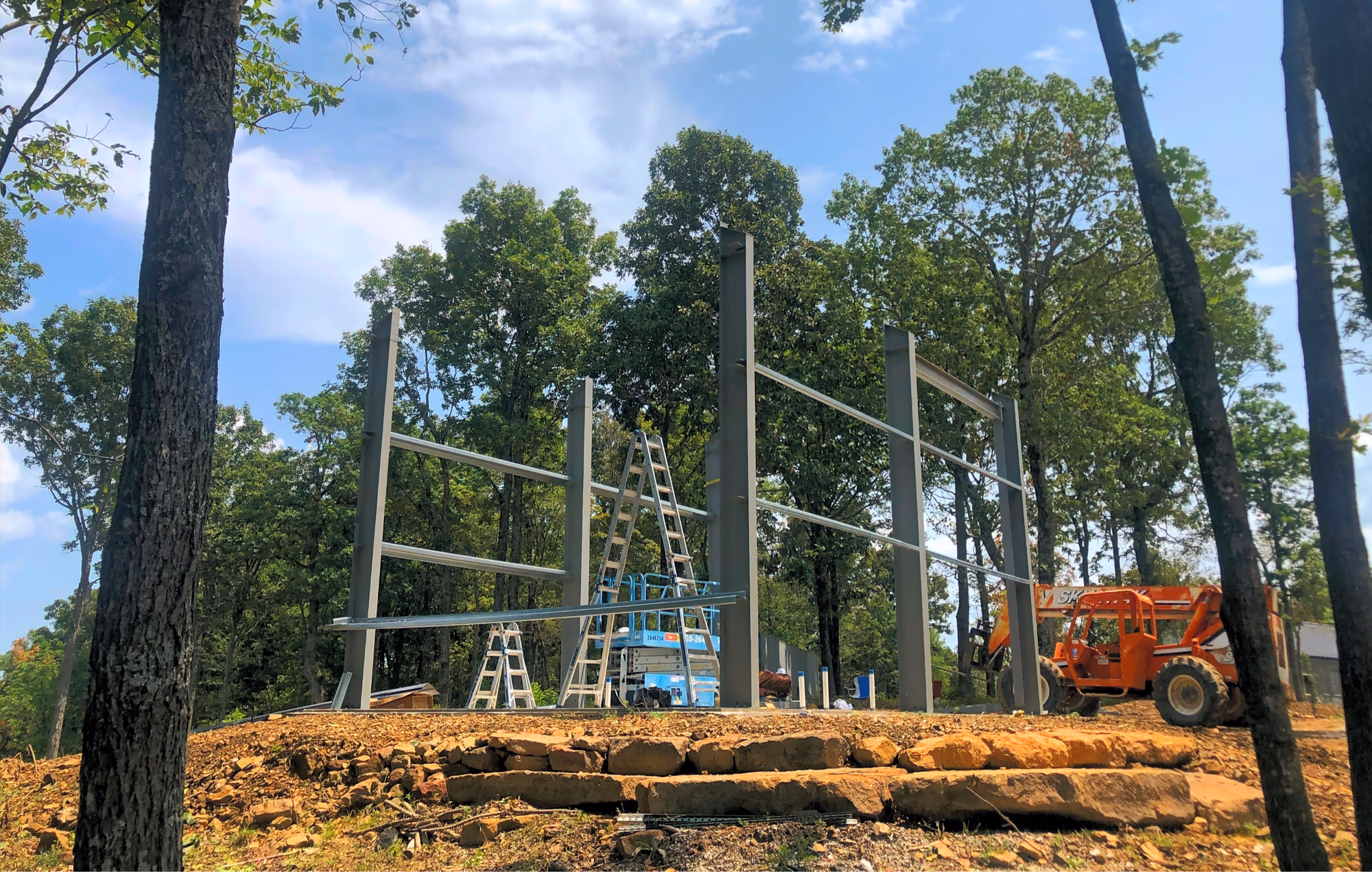 Red-iron columns and beams standing on a wooded hillside slab mid-erection, with a telehandler and scissor lift alongside