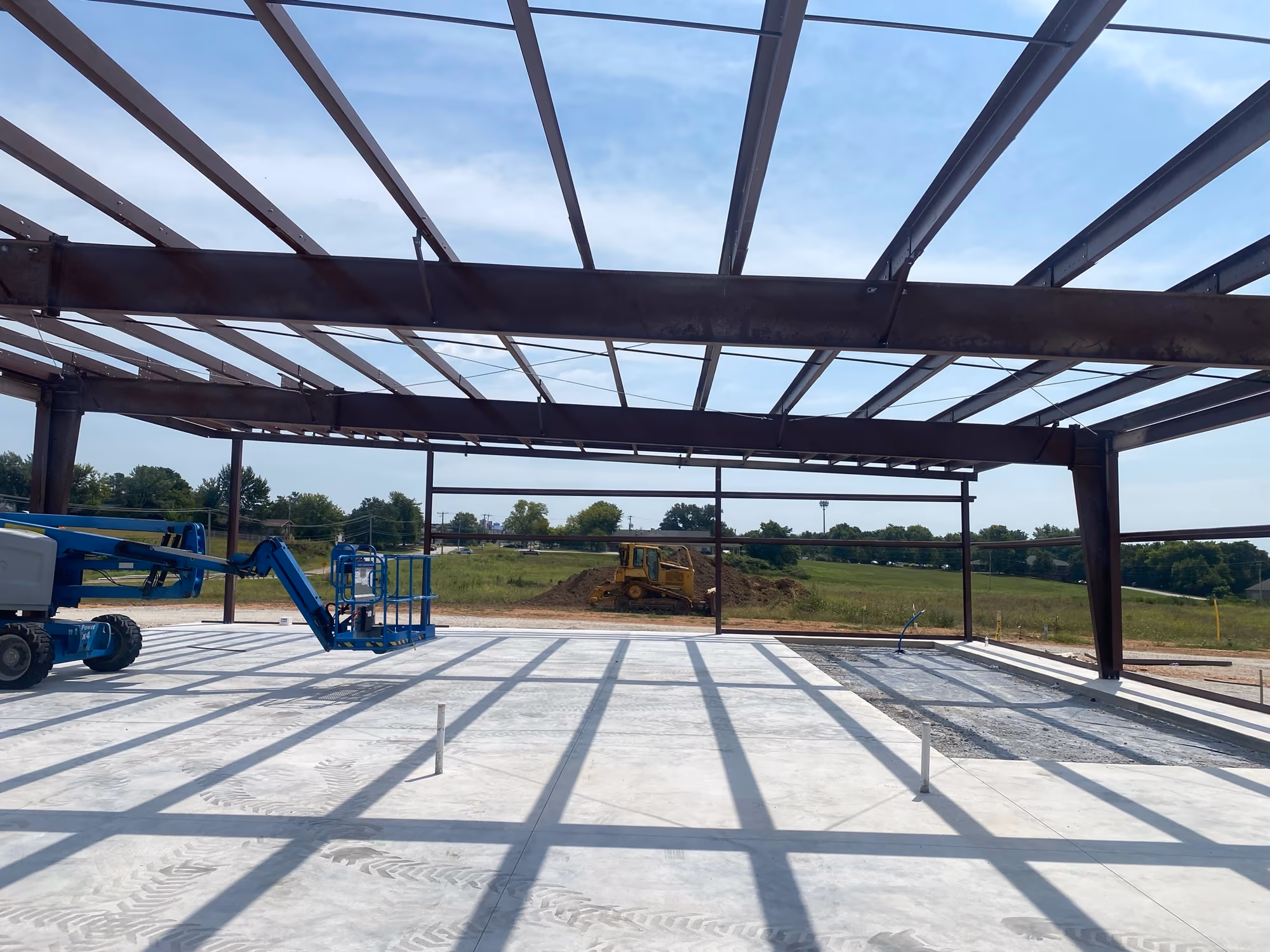 A red-iron steel frame standing on a finished concrete slab, with roof purlins in place and the building's footprint visible