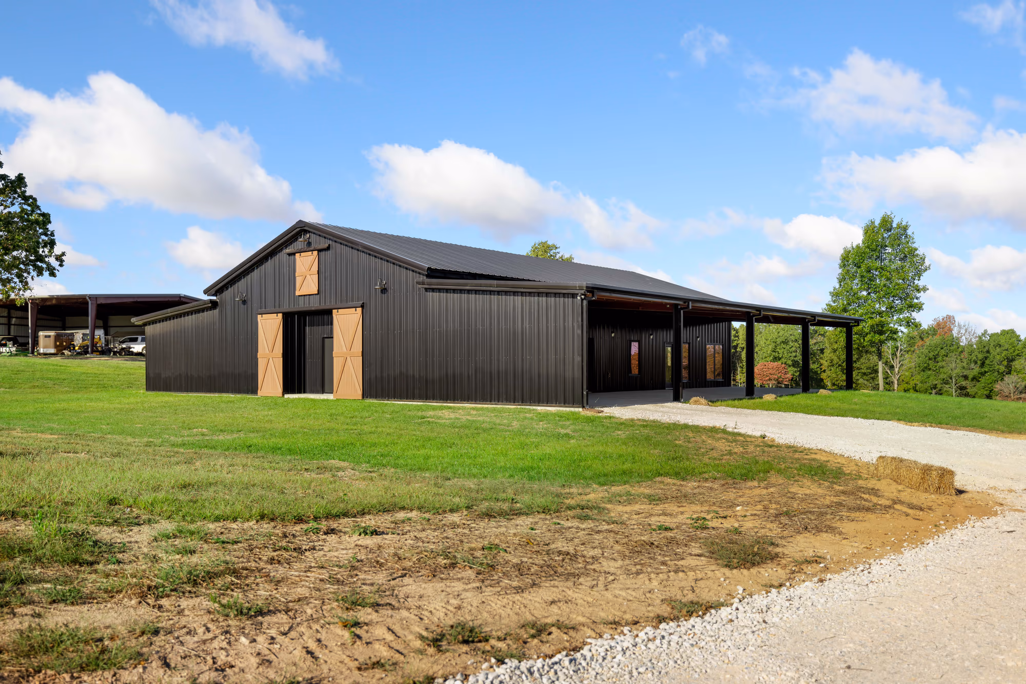 A black metal barndominium with tan sliding barn doors and a covered lean-to porch, on a mown rural lot