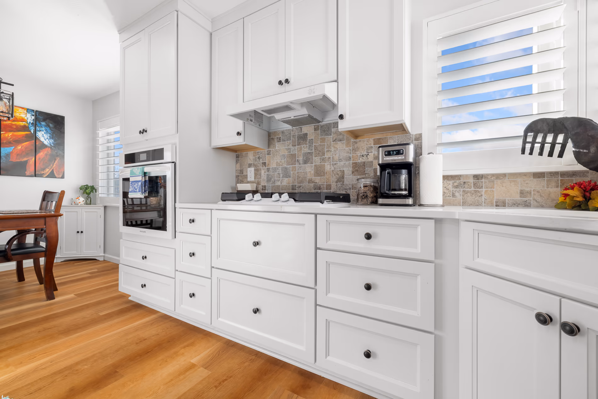 A finished kitchen in the Built Around the Hearth home — white shaker cabinetry and drawers with dark cup pulls, a travertine mosaic backsplash, a built-in wall oven, plantation shutters and an oak floor running through to the dining table
