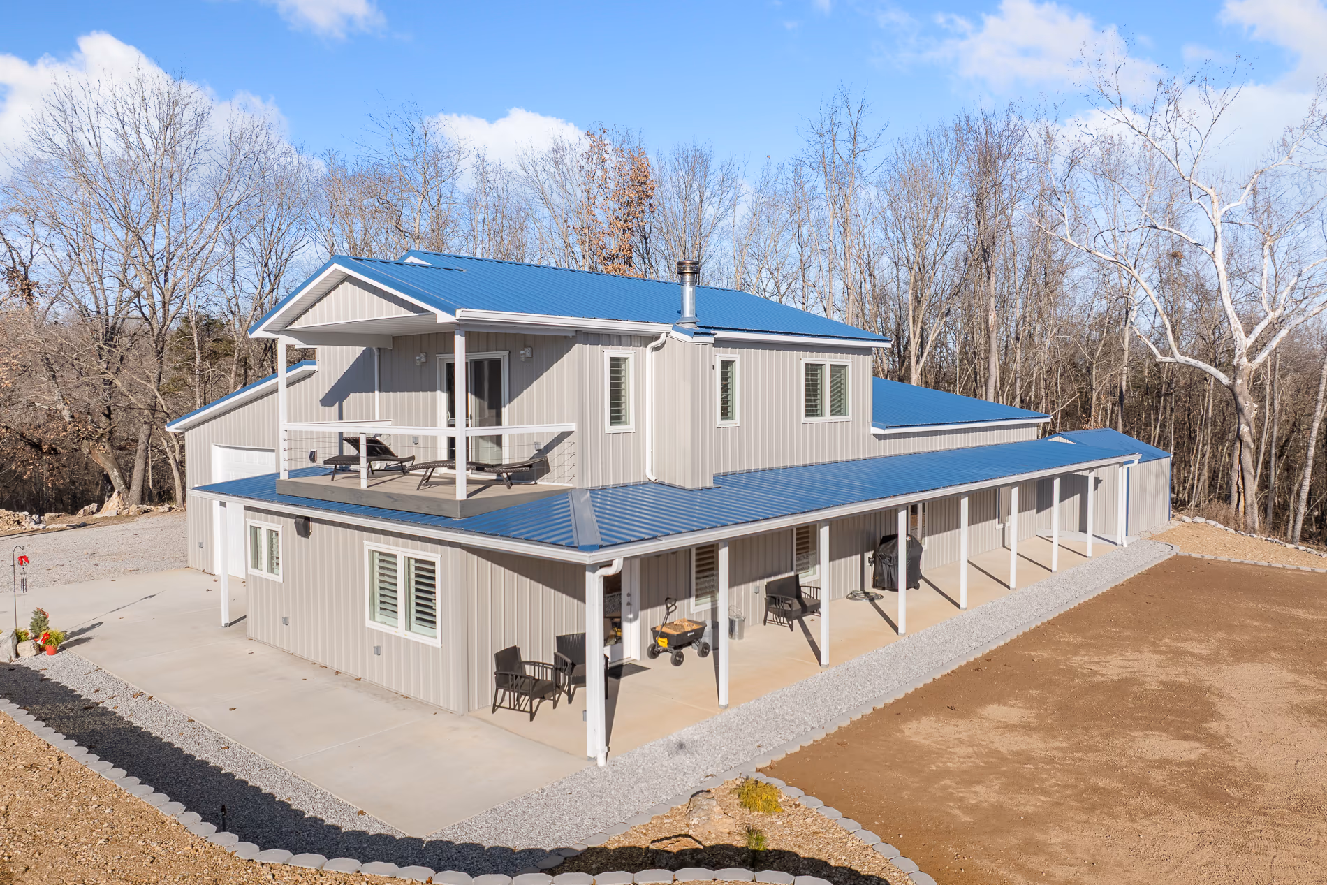 A two-storey grey home with a blue standing-seam metal roof, a wraparound covered porch and an upper deck, on a cleared lot in winter woods