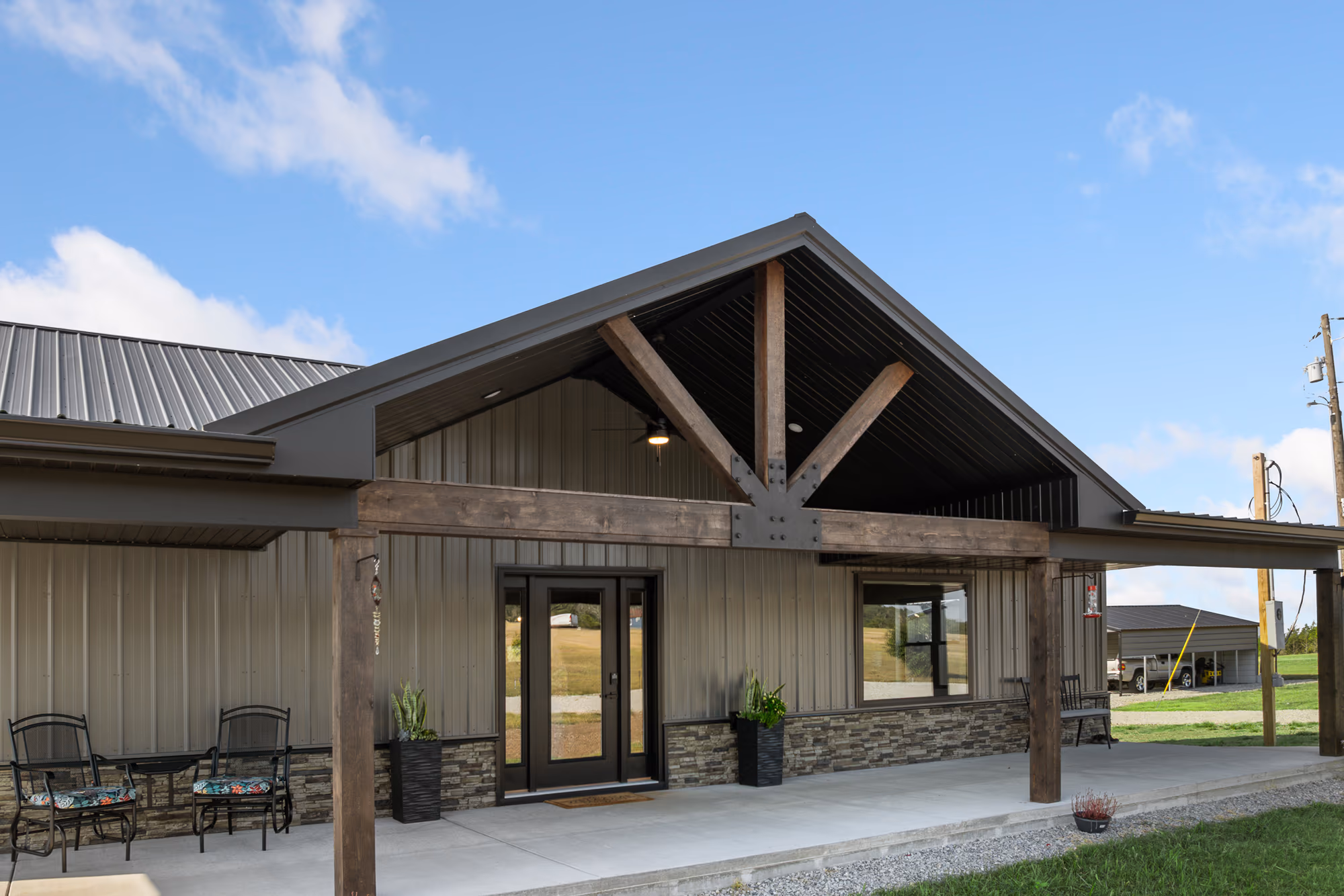 Covered front porch of a contemporary barndominium — stained timber trusses joined by a bolted steel plate, tan vertical metal siding and a stacked-stone wainscot