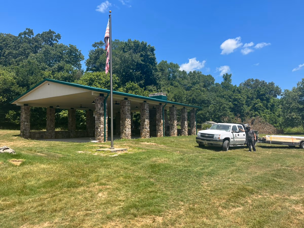 The Arkansas Game & Fish Pavilion after its re-roof — an open-air pavilion on stacked-stone columns with new white metal roofing, green trim, soffit and gutters