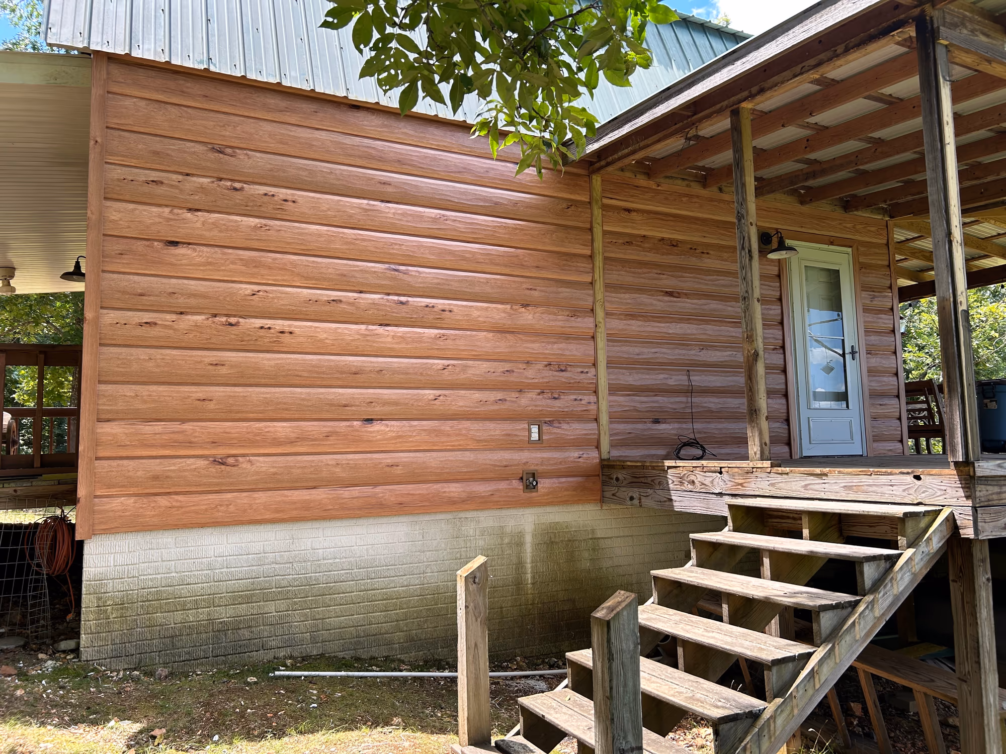 TrueLog™ metal siding installed on a cabin — horizontal steel panels with a printed wood grain and knots that read as stacked logs, running above a painted brick foundation, beside a treated timber porch and stairs under a green metal roof