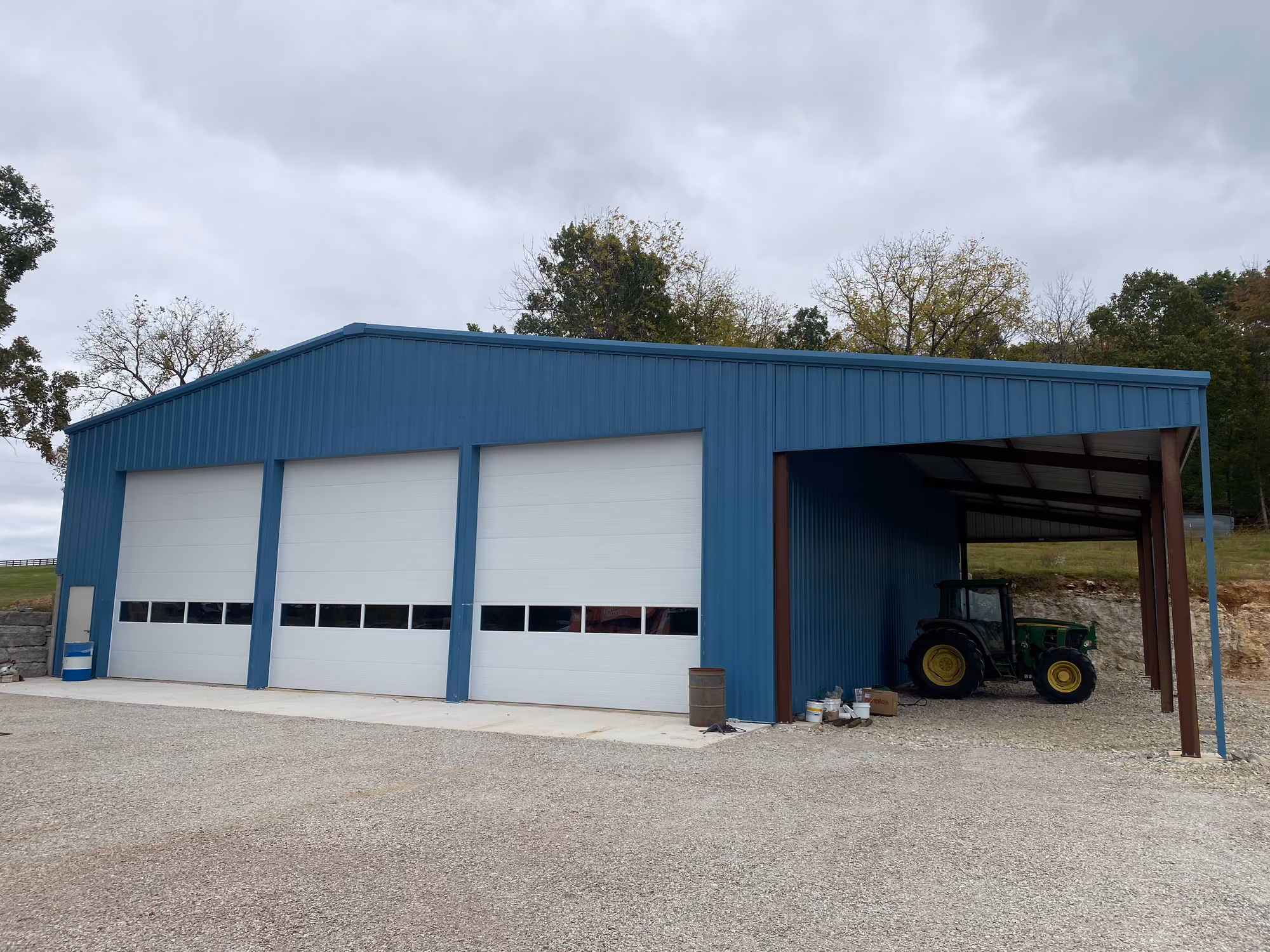A blue metal shop building with three white overhead doors and an open lean-to bay sheltering a tractor