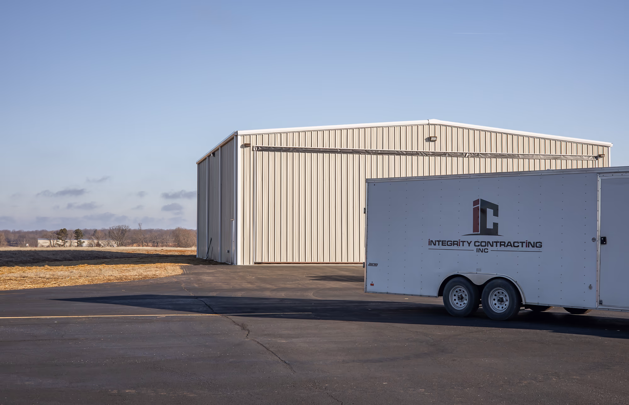 A cream steel commercial building with a full-width bifold door, an Integrity Contracting Inc box trailer parked on the apron in front of it