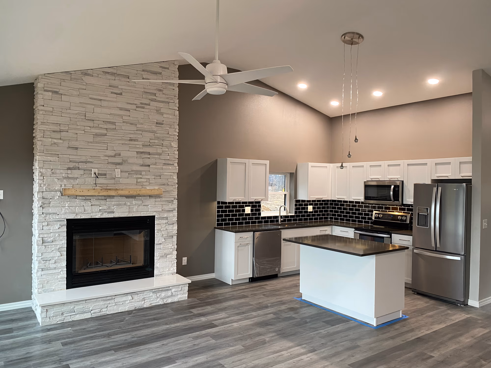 The open-concept interior of a finished barndominium — a floor-to-ceiling white stacked-stone fireplace with a raw timber mantel beside a vaulted-ceiling kitchen with white shaker cabinets, black subway tile and a centre island