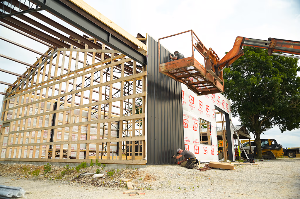 Two Integrity crew members hanging charcoal vertical metal siding over a steel frame and wood wall framing, one working from a boom lift and one at ground level