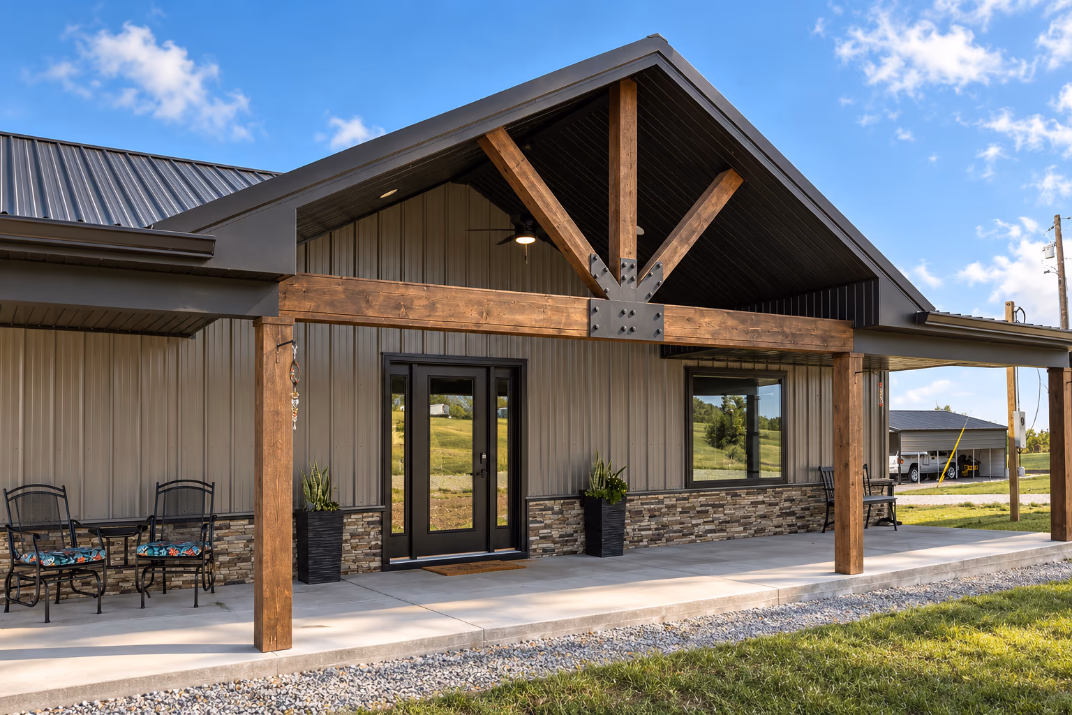 A custom home with a covered entry porch — a stained timber truss and bolted steel plate beneath a dark gable, tan vertical metal siding over a stacked-stone wainscot and a black-framed glass door